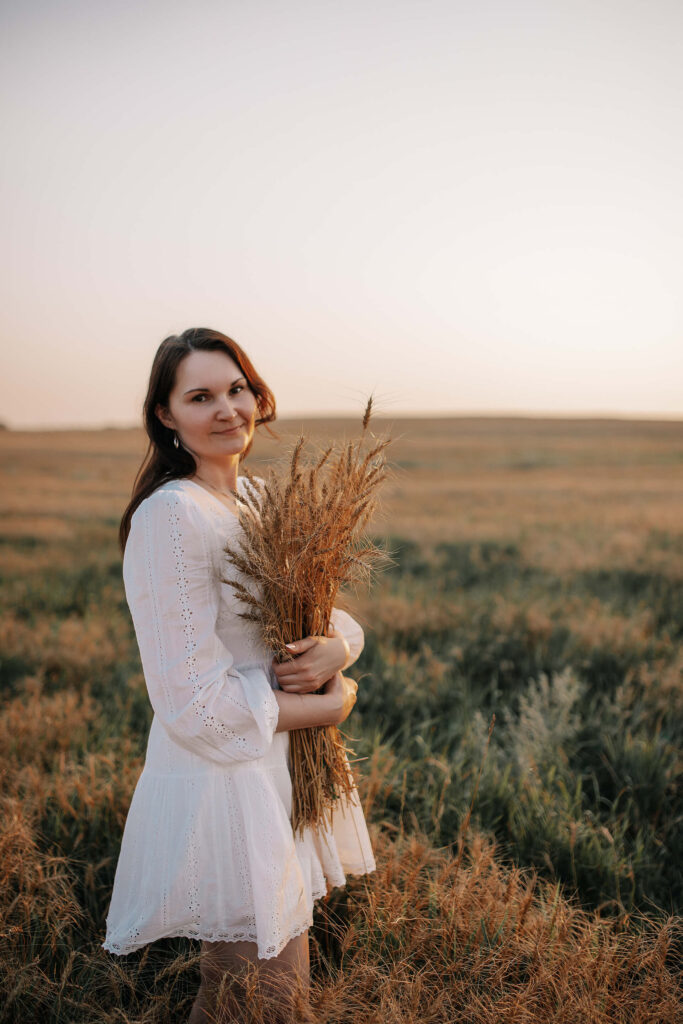 Tania in a white dress holding wheat in a field during sunset.