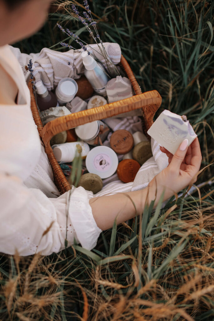 Tania seated in a field with a basket of natural cosmetics, holding up a signature soap bar.
