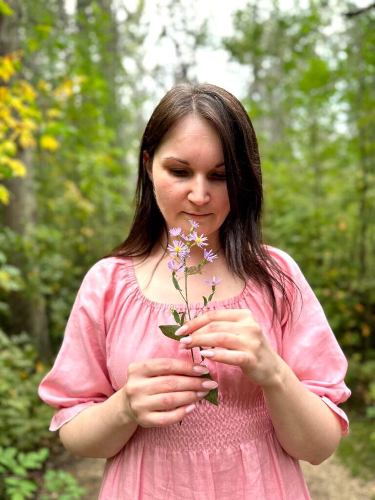 Tania in a soft pink dress holding and looking at a flower in the forest.